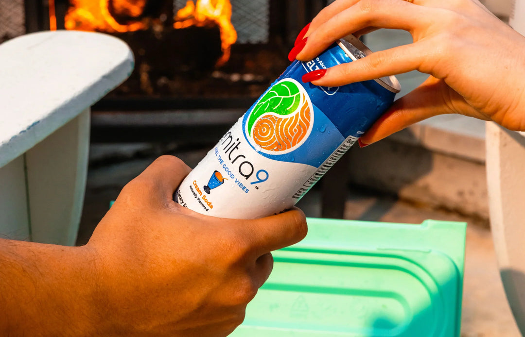 Mitra9 Cream Soda functional seltzer can being opened outdoors by a fire pit, lifestyle shot featuring a non-alcoholic functional drink with mitragynine and kava, refreshing beverage in hand
