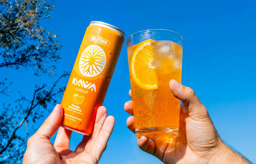 Hand holding a Mitra9 Kava Seltzer functional drink in Orange Dreamsicle flavor next to a refreshing mocktail with ice and fresh orange slice outdoors, showcasing a kava soda alternative to traditional cocktails