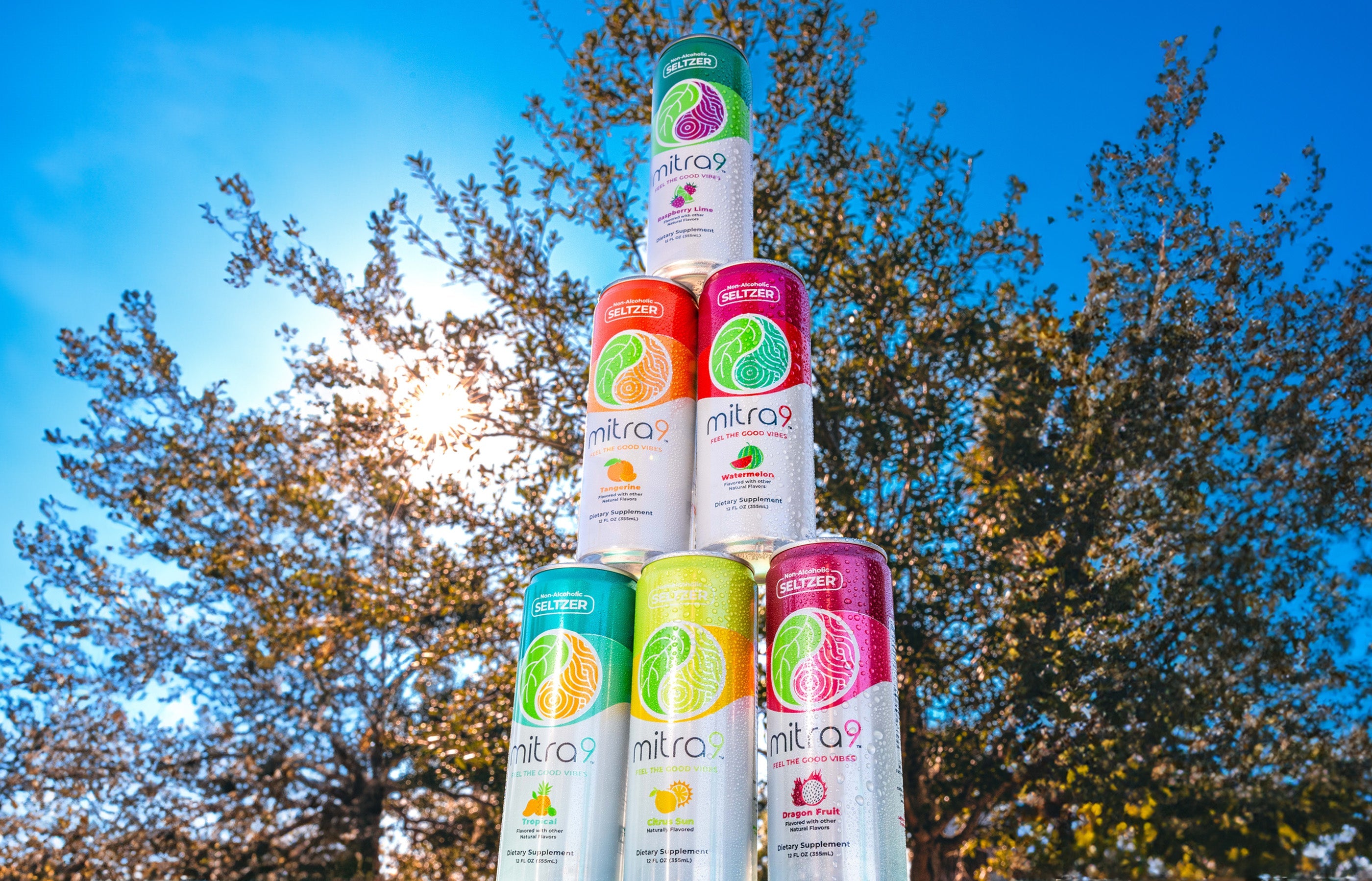 Stacked Mitra9 functional drink seltzers in multiple flavors outdoors with sunlight through trees, showcasing colorful non-alcoholic seltzer beverages for refreshing wellness moments