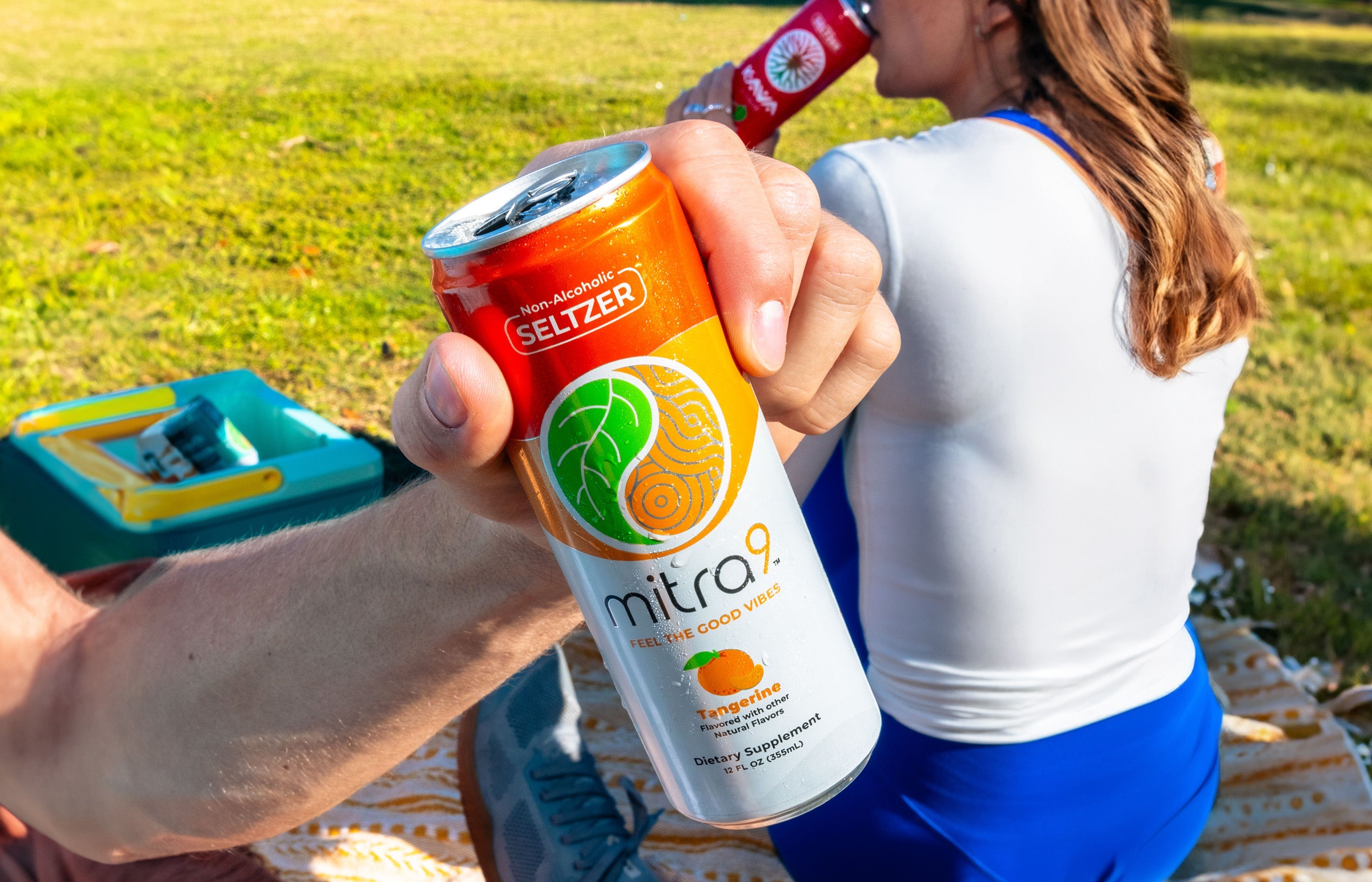 Hand holding a Mitra9 Tangerine functional drink seltzer outdoors while a person relaxes on a picnic blanket, showcasing a refreshing non-alcoholic seltzer alternative for social moments