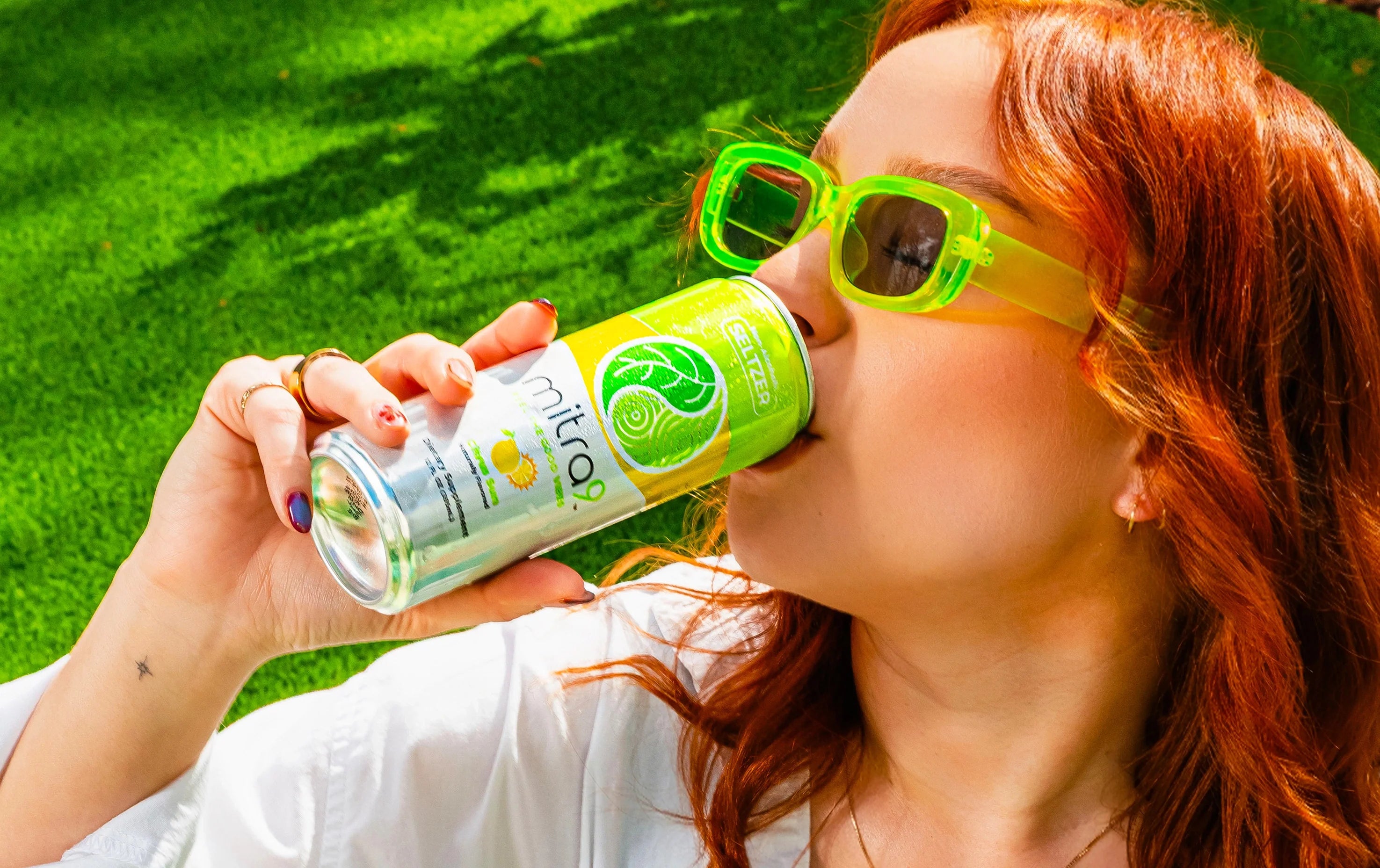 Woman wearing neon green sunglasses drinking Mitra9 Citrus Sun functional seltzer outdoors, refreshing kava soda and functional beverage lifestyle image