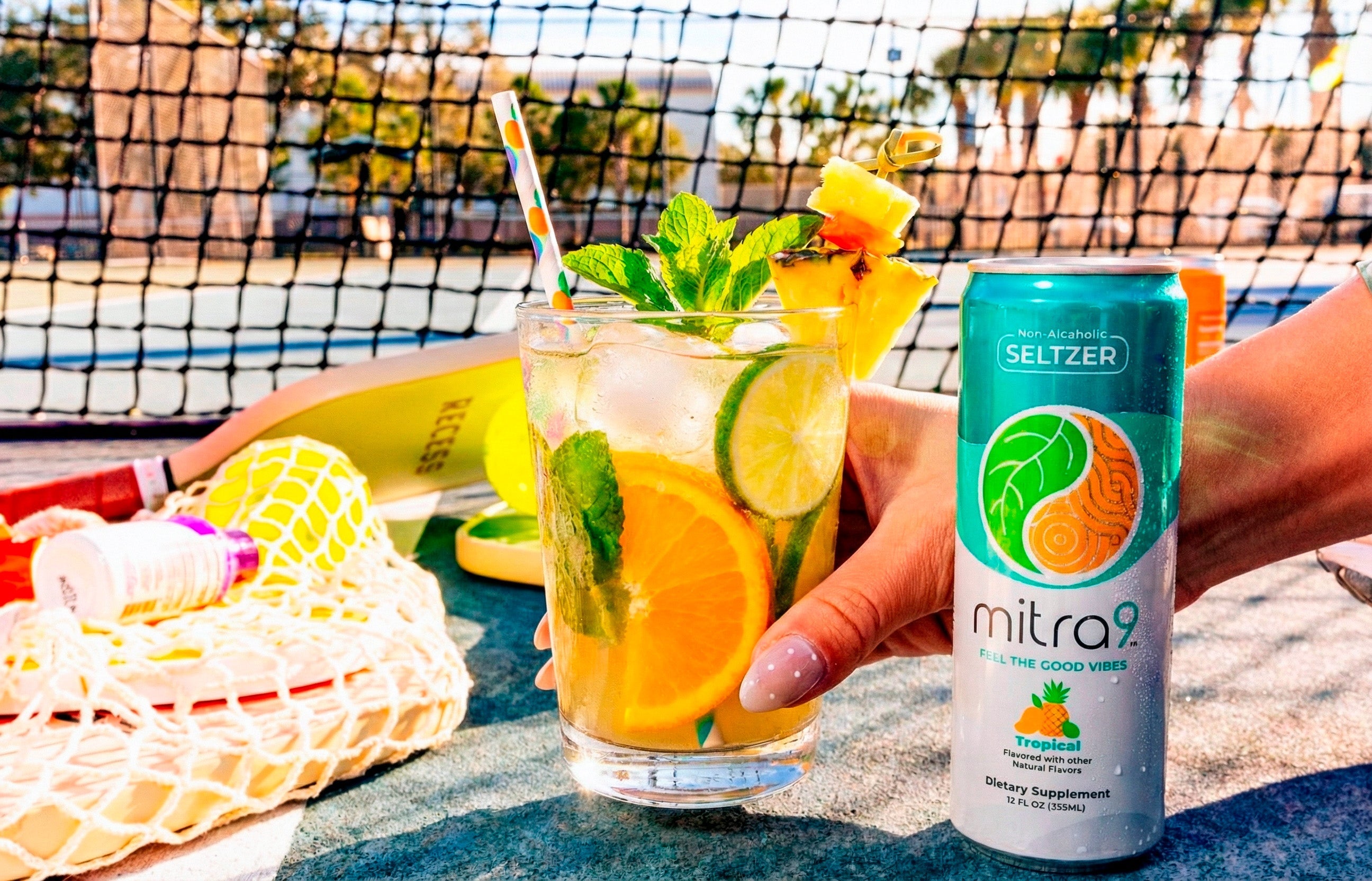 Lifestyle photo of Mitra9 functional drink tropical kratom seltzer can next to a fresh citrus mocktail with orange slices, lime, mint, and ice on a sunny outdoor court, showcasing a non-alcoholic wellness beverage for mood boost and relaxation