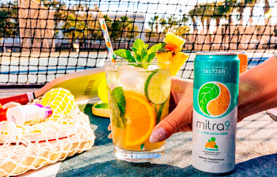 Lifestyle photo of Mitra9 functional drink tropical kratom seltzer can next to a fresh citrus mocktail with orange slices, lime, mint, and ice on a sunny outdoor court, showcasing a non-alcoholic wellness beverage for mood boost and relaxation