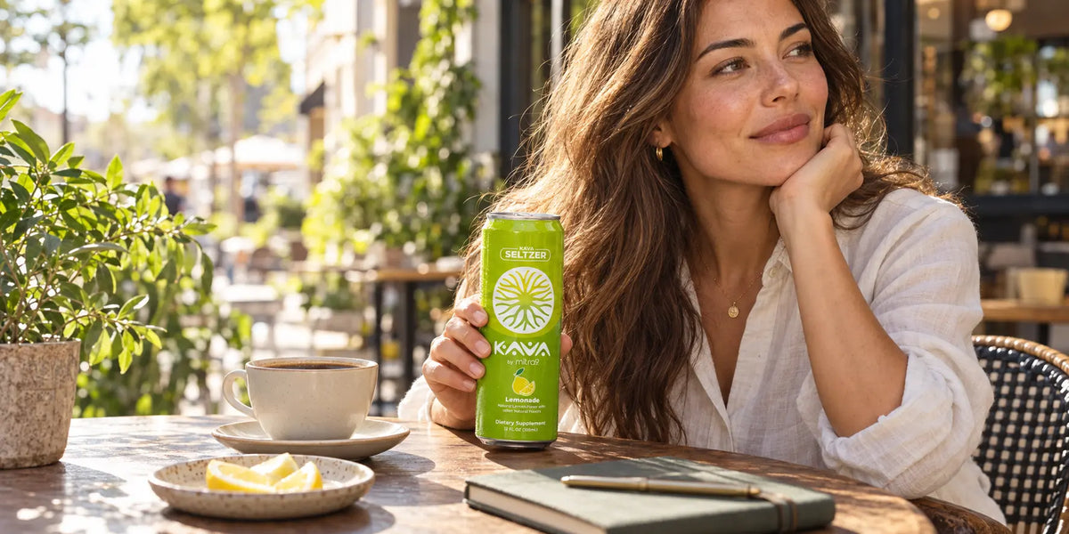Woman holding a Mitra9 Lemonade Kava Seltzer beside a cup of coffee at an outdoor café, illustrating kava and caffeine.