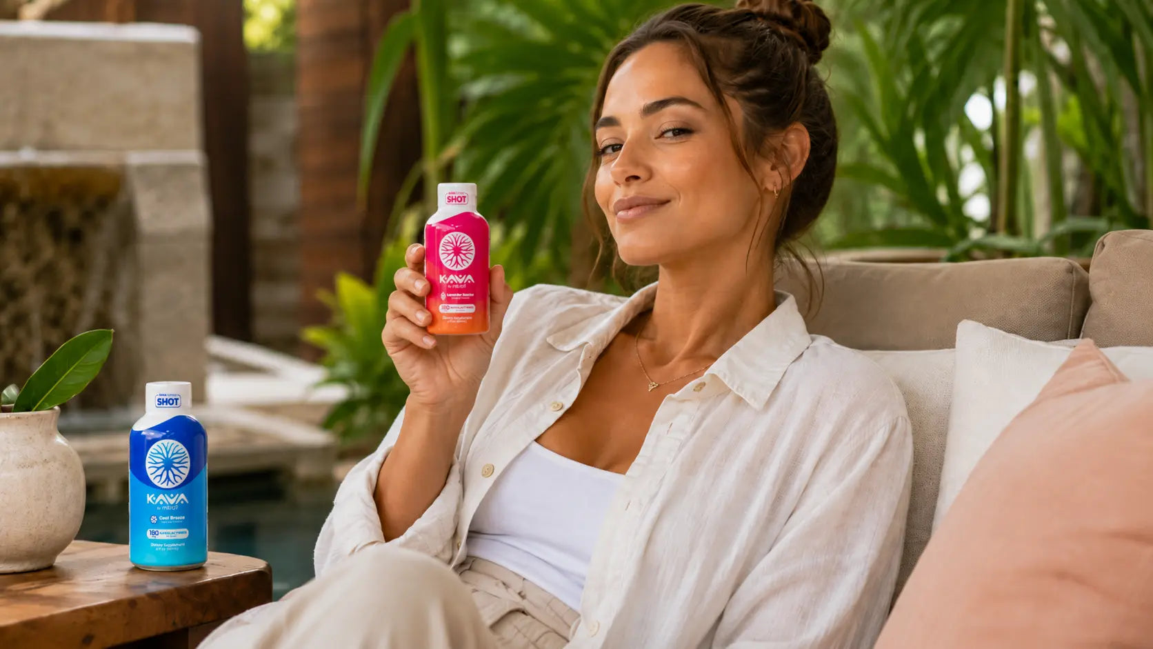 Woman presenting a Kava by Mitra9 Lavender Sunrise kava shot in a relaxing tropical setting with a Cool Breeze kava shot nearby.