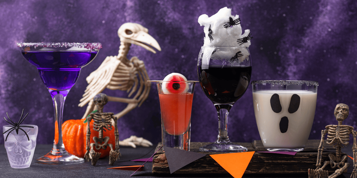 A variety of Halloween cocktails and mocktails with spooky decorations, including skeletons, an eyeball garnish, cotton spider webs, and themed glasses.