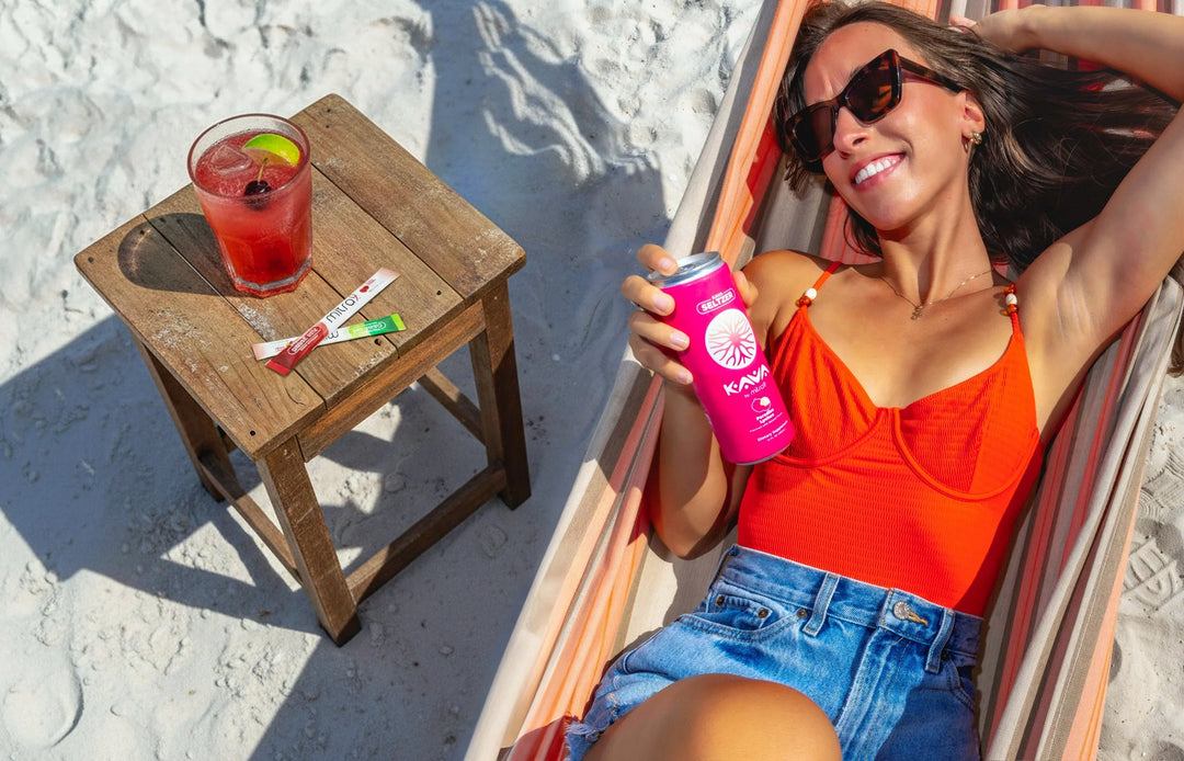 Woman relaxing on a beach hammock while holding a Mitra9 kava seltzer can beside a tropical drink on a wooden table