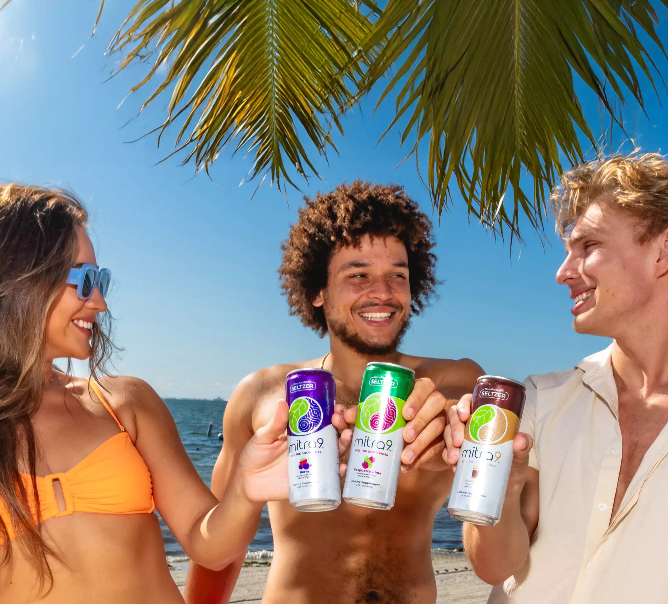 Three people on a beach holding drinks with palm leaves and blue sky in the background